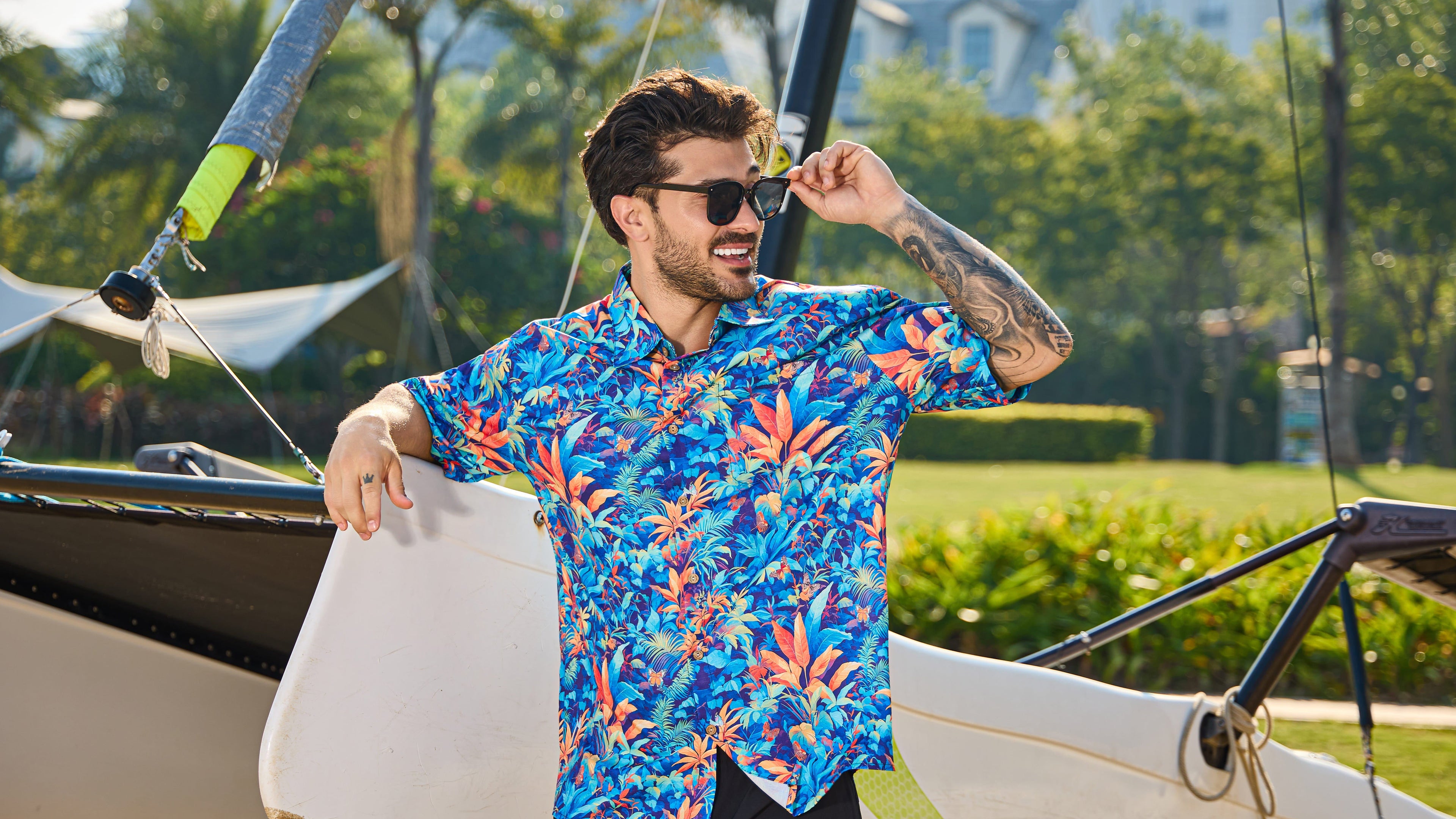 Man in a colorful shirt standing next to a sailboat with trees and grass in the background