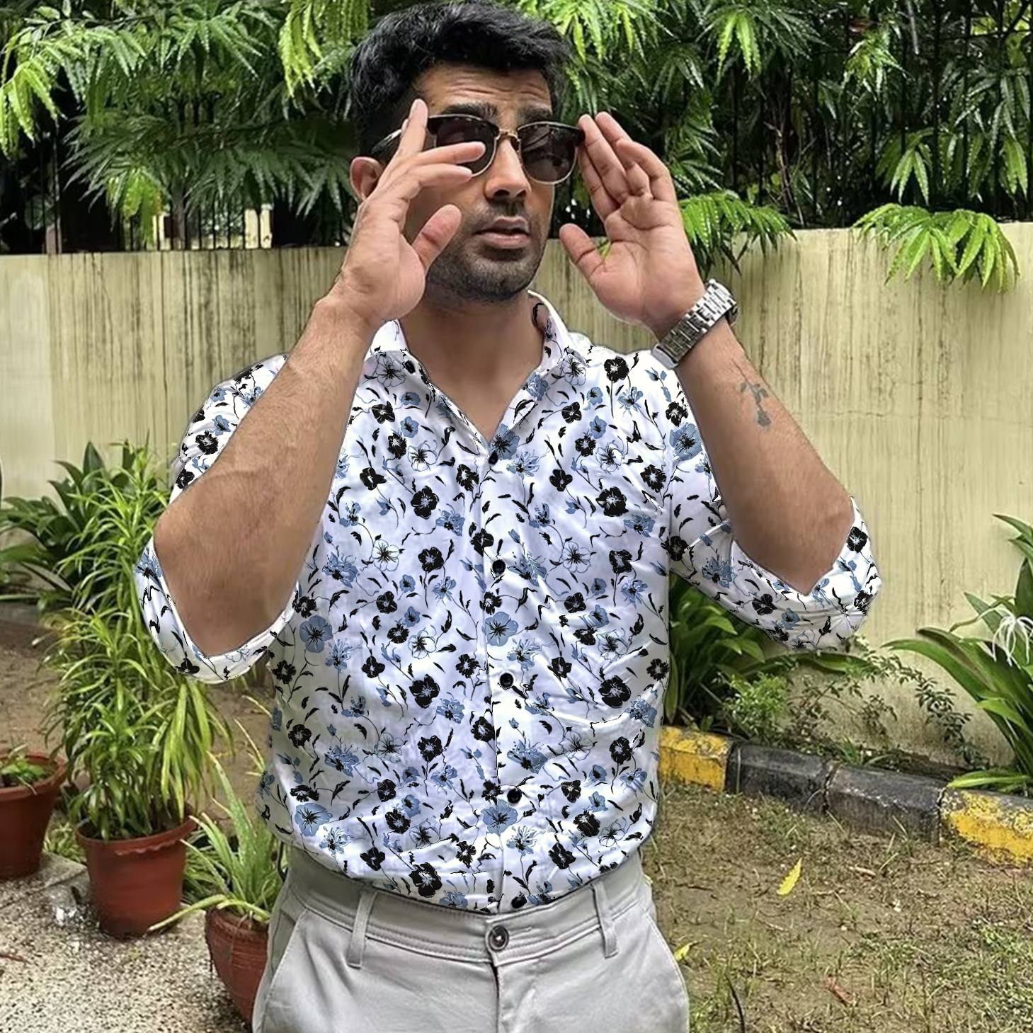 A man wearing a dark-colored shirt featuring a beautiful pattern of white and blue flowers, standing confidently outdoors with sunglasses on.