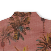 Close-up of the back collar area of a Hawaiian shirt, highlighting a reddish fabric with a palm tree design in earthy tones of brown, green, and yellow, giving it a vintage tropical feel. The pattern is slightly faded, adding to the casual, sun-bleached aesthetic.