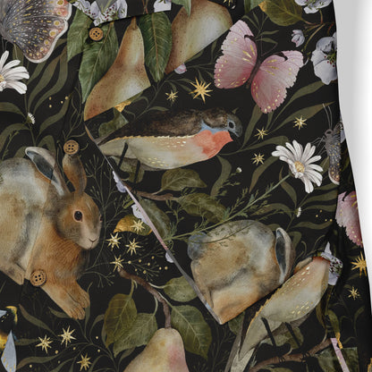 Close-up view of a shirt featuring a whimsical design with a rabbit, birds, pears, butterflies, and flowers against a dark background, accented with small golden stars.