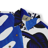 This is a close-up of a shirt featuring a bold, abstract design with large, vibrant blue and black leaves set against a white background. The shirt includes wooden buttons and the Kalvero label, adding a natural touch to the dynamic and eye-catching pattern.