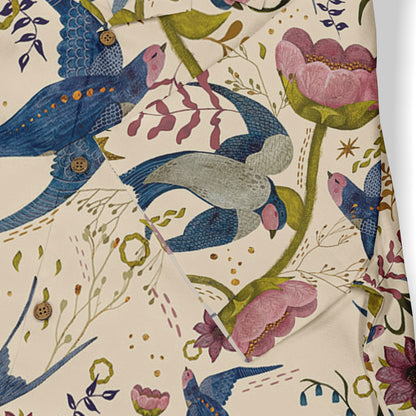 This is a close-up of a shirt featuring a whimsical design with vibrant blue birds and pink flowers, set against a light beige background. The detailed pattern includes floral elements and stars, along with wooden buttons, enhancing the playful and artistic nature of the shirt.